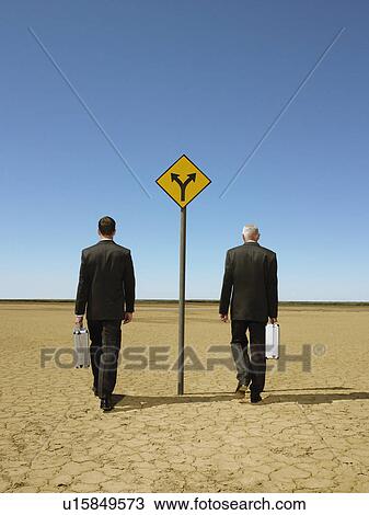 Two businessmen with briefcases walking past road sign in desert back view View Large Photo Image Stock Image - Two businessmen with briefcases walking past road sign in desert back view. Fotosearch