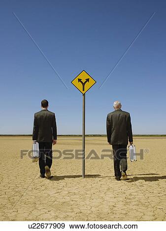 Two businessmen with briefcases walking past road sign in desert back view View Large Photo Image Stock Photo - Two businessmen with briefcases walking past road sign in desert back view. Fotosearch