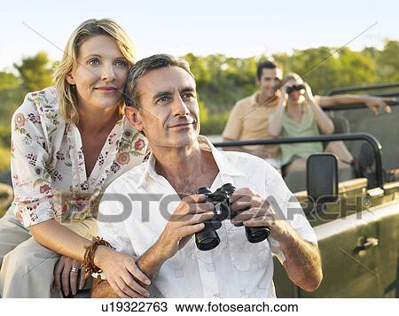 Stock Image - Two couples on trip one sitting in jeep using binoculars. Fotosearch