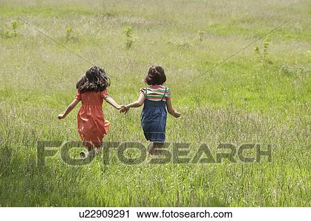 Stock Image - Two girls (7-9) holding hands running in field. Fotosearch
