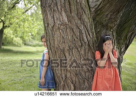Stock Image - Two girls (7-9) playing hide and seek by tree. Fotosearch