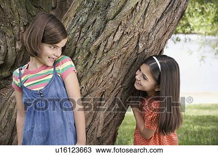 Stock Image - Two girls (7-9) playing hide and seek by tree. Fotosearch