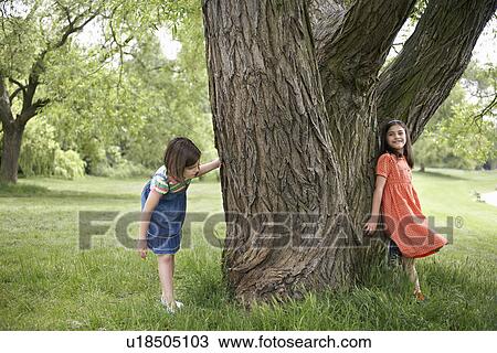 Stock Image - Two girls (7-9) playing hide and seek by tree. Fotosearch