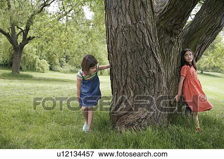 Stock Photo - Two girls (7-9) playing hide and seek by tree. Fotosearch