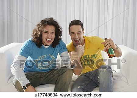 Picture - Two men sitting on sofa watching television and cheering with beer bottle. Fotosearch