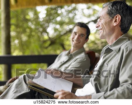 Stock Photography - Two men sitting on terrace smiling one holding book. Fotosearch