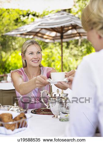 Stock Photography - Two women sitting at table passing tea cup selective focus. Fotosearch