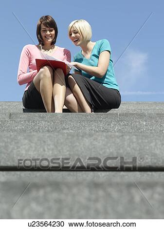 Stock Image - Two women sitting on stairs outdoors reading papers and laughing low angle view. Fotosearch