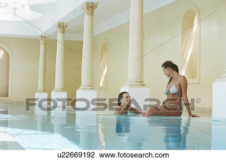 Stock Image - Two young women talking at edge of swimming pool. Fotosearch