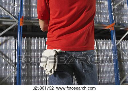 Stock Image - Warehouse Worker standing in front of full shelves gloves hanging from back pocket mid section rear view. Fotosearch