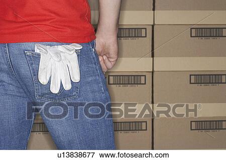 Stock Photo - Warehouse Worker standing in front of stacks of boxes gloves hanging out of back pocket mid section back view. Fotosearch