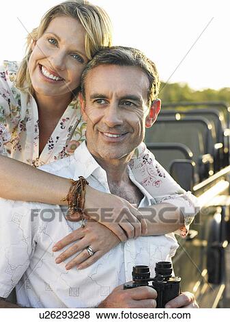 Stock Photo - Woman embracing man man holding binoculars jeep in background. Fotosearch