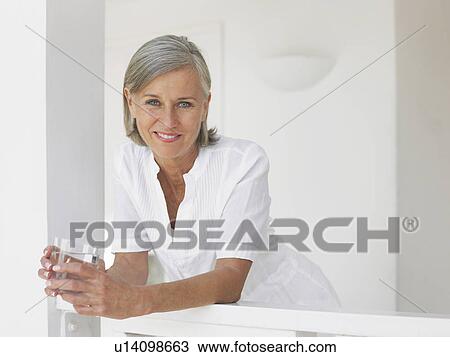 Stock Image - Woman holding glass of water leaning on verandah balustrade portrait. Fotosearch