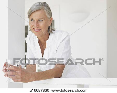 Stock Image - Woman holding glass of water leaning on verandah balustrade portrait. Fotosearch