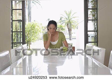 Stock Image - Woman reading book sitting at dining table. Fotosearch