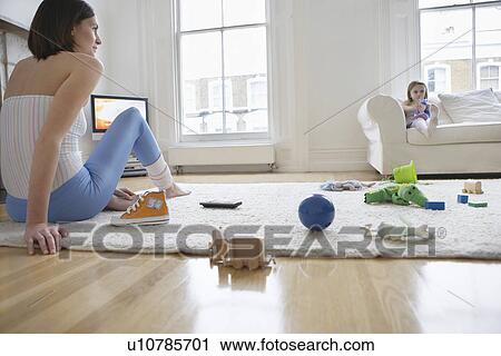 Stock Image - Woman sitting on floor surrounded with toys daughter (5-6) on sofa. Fotosearch