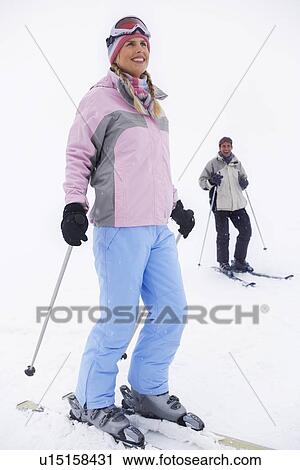 Stock Image - Woman skier standing on ski slope man on skis in background. Fotosearch