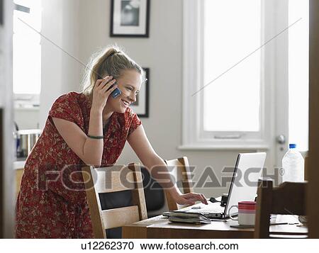 Stock Image - Woman using mobile phone while using laptop in dining room side view. Fotosearch