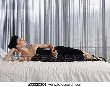 Picture - Woman wearing elegant dress lying on bed with champagne glass in bedroom. Fotosearch