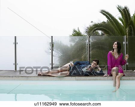 Stock Photo - Women reclining with champagne by outdoor pool. Fotosearch