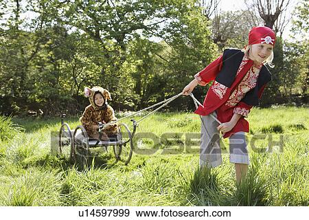 Stock Photo - Young boy (7-9) in pirate costume pulling boy (5-6) in jaguar costume sitting on cart smiling. Fotosearch
