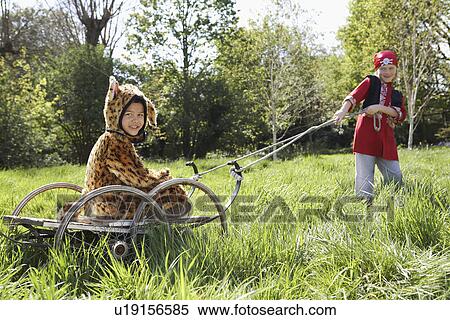 Stock Photography - Young boy (7-9) in pirate costume pulling boy (5-6) in jaguar costume sitting on cart smiling. Fotosearch