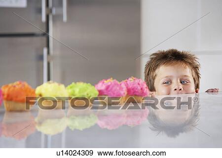Stock Photo - Young boy peeking over counter at row of cupcakes in kitchen. Fotosearch