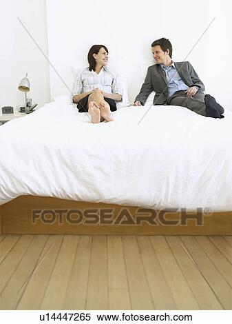 Stock Photography - Young business couple sitting on bed looking in eyes and smiling. Fotosearch