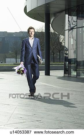 Stock Photo - Young business man holding bunch of flowers and newspaper walking in street. Fotosearch