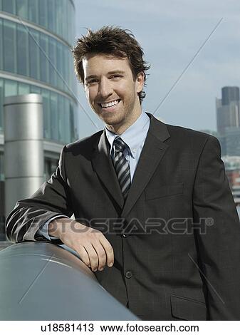 Stock Image - Young businessman leaning on pipe outside building smiling. Fotosearch