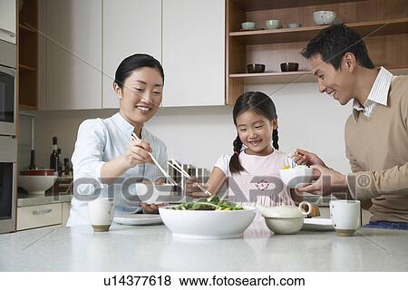 Stock Photo - Young couple and daughter eating a meal with chopsticks on kitchen counter. Fotosearch