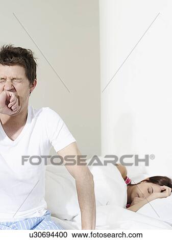 Stock Image - Young couple in bedroom woman sleeping man sitting on bed and yawning. Fotosearch