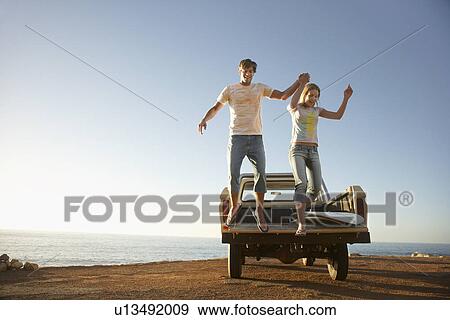 Stock Photo - Young couple jumping from back of van parked by ocean. Fotosearch