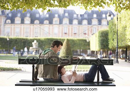 Stock Image - Young couple reading book sitting on bench in park. Fotosearch