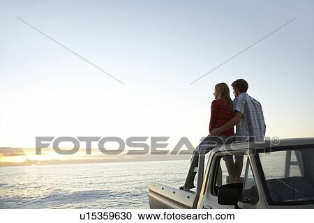 Stock Image - Young couple sitting on van parked in front of ocean. Fotosearch