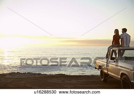 Stock Image - Young couple sitting on van parked in front of ocean. Fotosearch