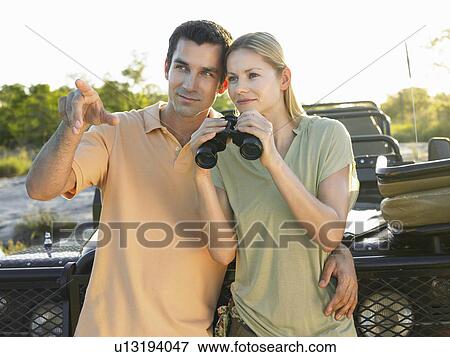 Young couple standing by jeep man pointing woman holding binoculars View Large Photo Image Stock Photo - Young couple standing by jeep man pointing woman holding binoculars. Fotosearch