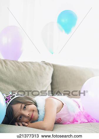 Stock Image - Young girl (7-9) lying on sofa with balloons. Fotosearch