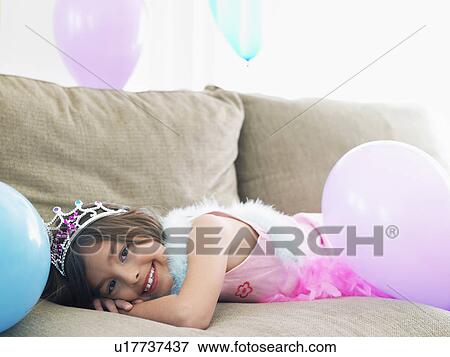 Stock Photo - Young girl (7-9) lying on sofa with balloons smiling. Fotosearch