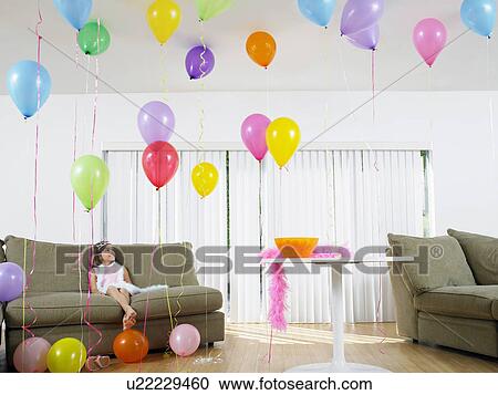 Stock Image - Young girl (7-9) sitting in living room full of balloons. Fotosearch