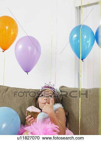 Stock Image - Young girl (7-9) sitting on sofa eating cupcake looking up at balloons. Fotosearch