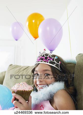 Stock Photograph - Young girl (7-9) sitting on sofa eating cupcake close-up. Fotosearch