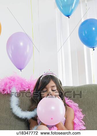 Stock Image - Young girl (7-9) sitting on sofa blowing up balloon. Fotosearch