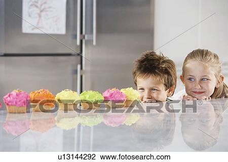 Young girl and boy looking over counter at row of cupcakes in kitchen View Large Photo Image Stock Image - Young girl and boy looking over counter at row of cupcakes in kitchen. Fotosearch