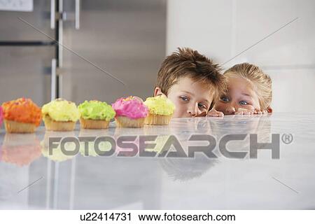 Stock Image - Young girl and boy peeking over counter at row of cupcakes high section. Fotosearch