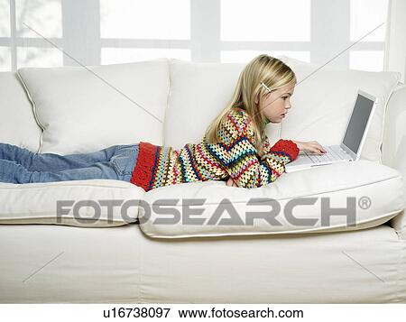 Stock Photo - Young Girl lying on stomach on sofa using Laptop side view. Fotosearch