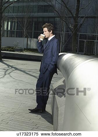 Stock Photography - Young man in suit leaning on pipe outdoors drinking coffee side view. Fotosearch
