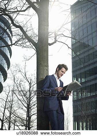 Stock Photo - Young man in suit standing under tree outside office building reading newspaper. Fotosearch