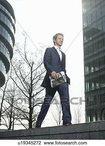 Stock Image - Young man in suit walking on wall outside office building holding newspaper. Fotosearch