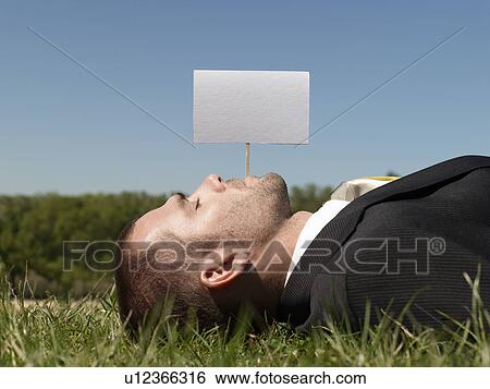 Stock Photograph - Young man lying on grass by blank sign. Fotosearch